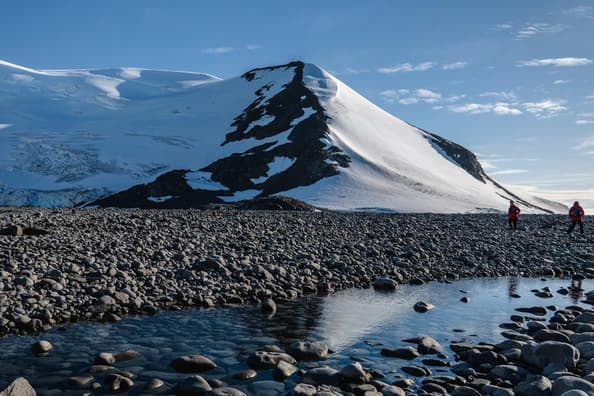 Two people in red jackets walking on a rocky Antarctic beach.
