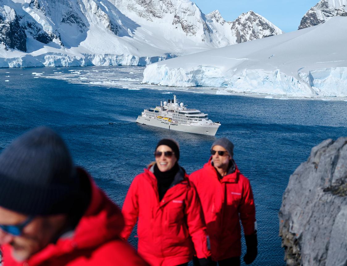 People hiking in red jackets with an expedition ship and snow-covered mountains in Antarctica.