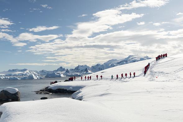 People in red jackets hiking up a snowy Antarctic hill with icebergs and mountains in the background.