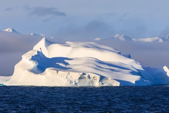 A large iceberg floating in the ocean with snowy mountains in the background.